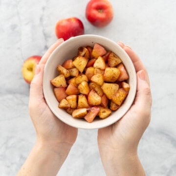 A bowl of cubed cooked cinnamon spiced apples being held above a marble bench top.