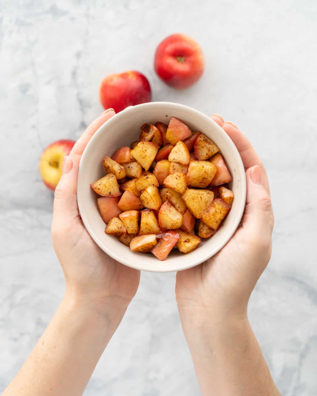 A bowl of cubed cooked cinnamon spiced apples being held above a marble bench top. 