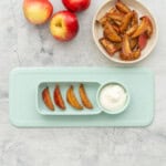 A silicone baby plate with four slices of cooked apples and a dipping bowl of yogurt on a bench next to a plate of more apple slices.
