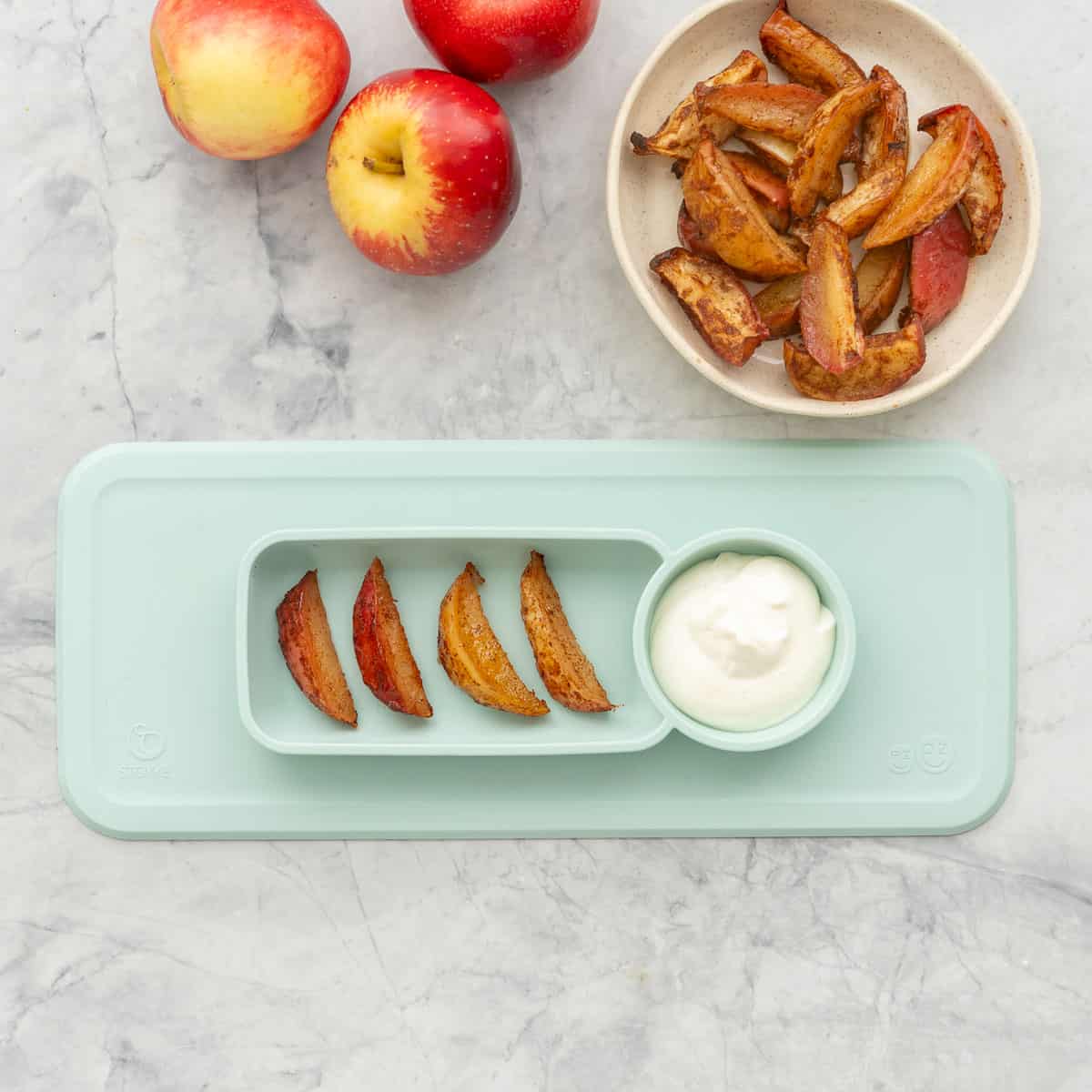 A silicone baby plate with four slices of cooked apples and a dipping bowl of yogurt on a bench next to a plate of more apple slices. 
