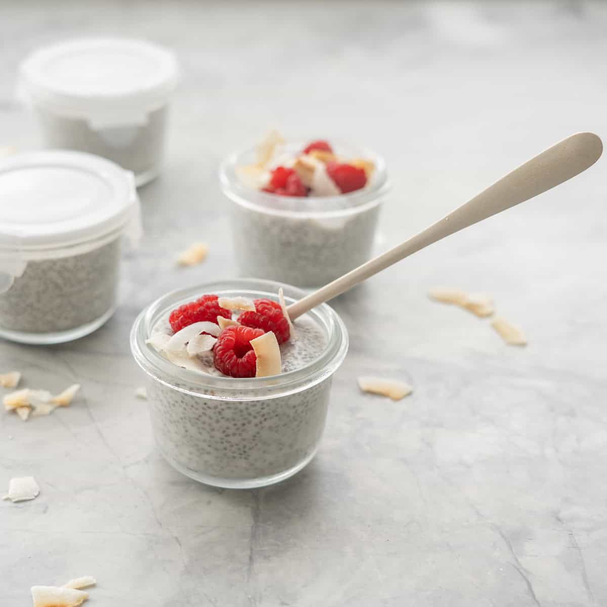 A small glass container filled with chia seed pudding, topped with raspberries coconut wood spoon in bowl.