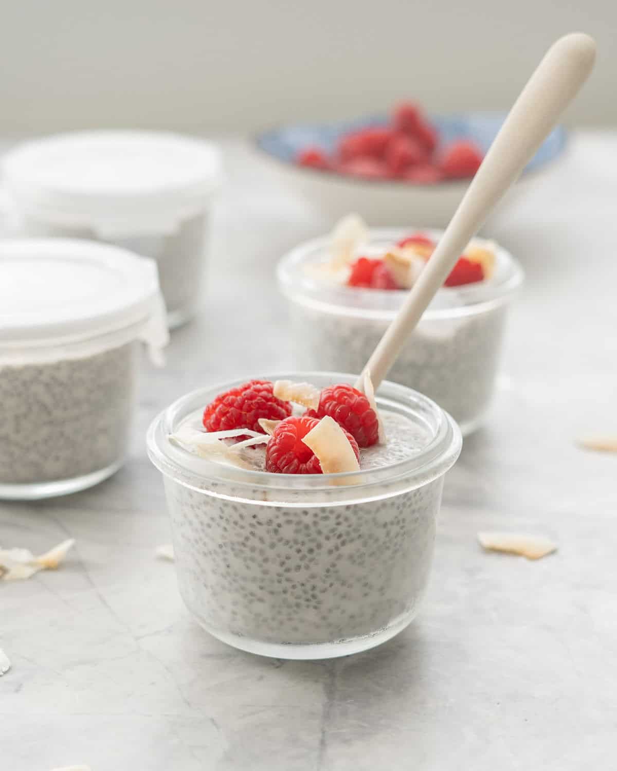 A small glass container of coconut chia pudding topped with raspberries and coconut flakes, wood spoon in bowl.
