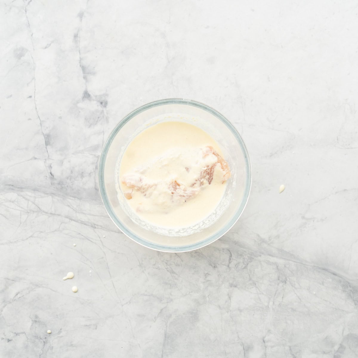 A chicken breast submerged in a white slurry in the bottom, of a glass mixing bowl.