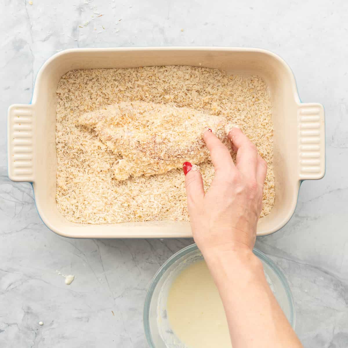 A chicken fillet being crumbed in an oblong baking dish filled with bread crumbs. 