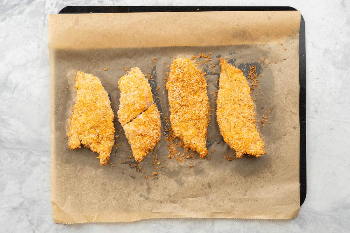 Four golden baked crumbed chicken cutlets on a lined baking sheet. 