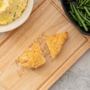 Looking down on a crumbed chicken cutlet that has been cut in half on a wooden chopping board.