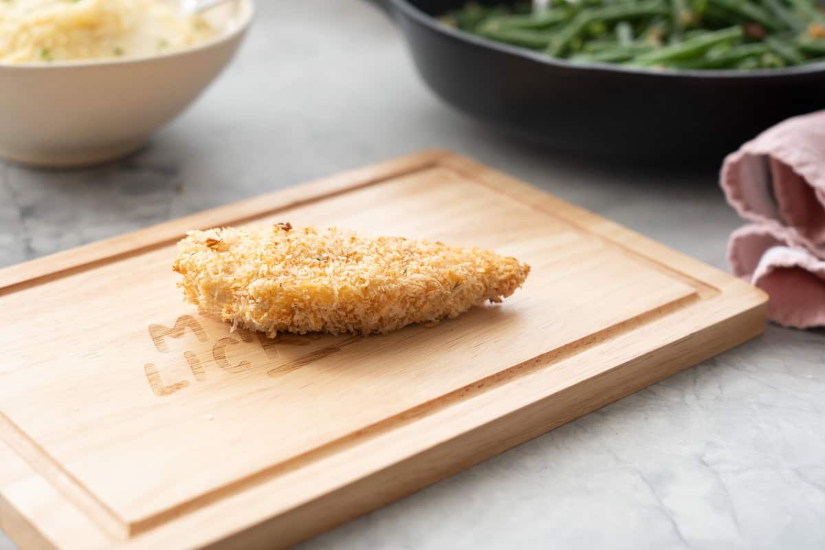 A crumbed baked chicken cutlet on a wooden chopping board in front of a bowl of beans.