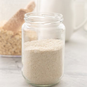 One jar of oat flour resting on the bench in front of a container of oats.