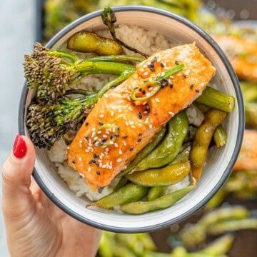 A bowl with green vegetables and a glazed piece of salmon sprinkled with sesame seeds being held above a tray of salmon and vegetables.