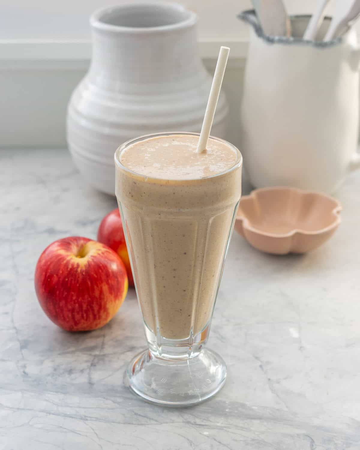 A tall sundae glass filled with a brown smoothie and a white paper straw, two red apples in the background.