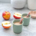 Two small green toddler cups with pink silicone straws filled with pale brown smoothie, apple pieces in the background.