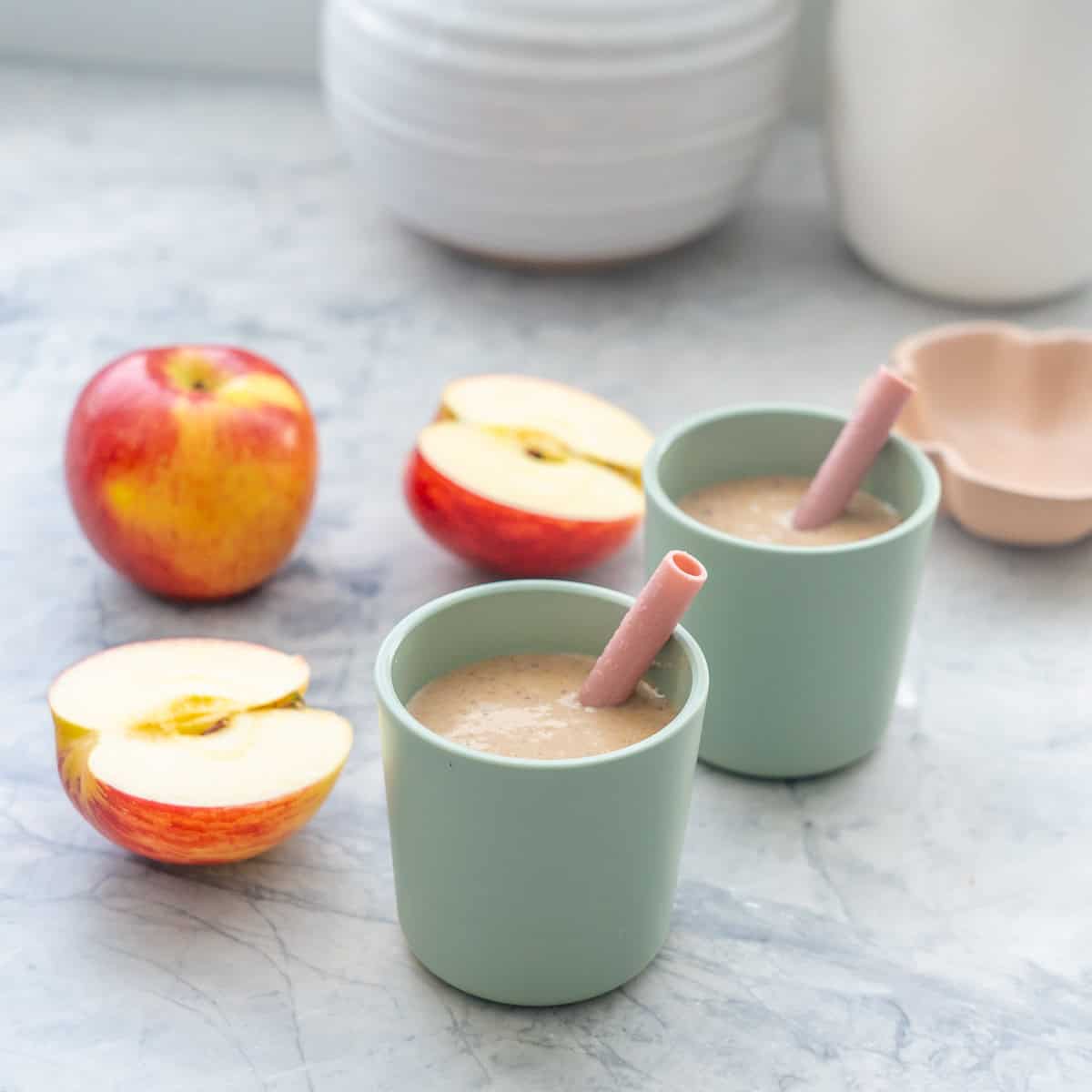 Two small green toddler cups with pink silicone straws filled with pale brown smoothie, apple pieces in the background.