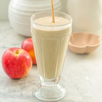 A tall sundae glass almost fill with smoothie, a thin stream of smoothie pouring in still visible, two apples and white ceramic jug behind the sundae glass.