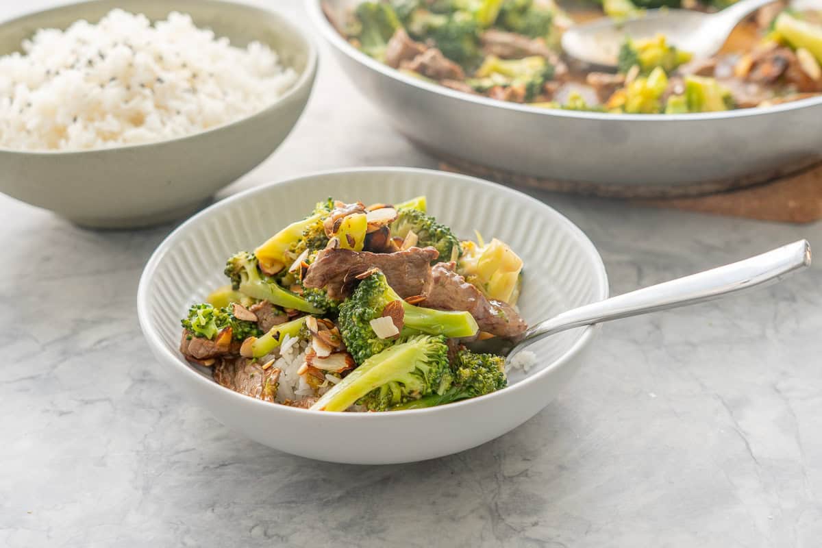 Broccoli beef dish served in dinner bowl with fork on rice garnished with roasted sliced almonds.