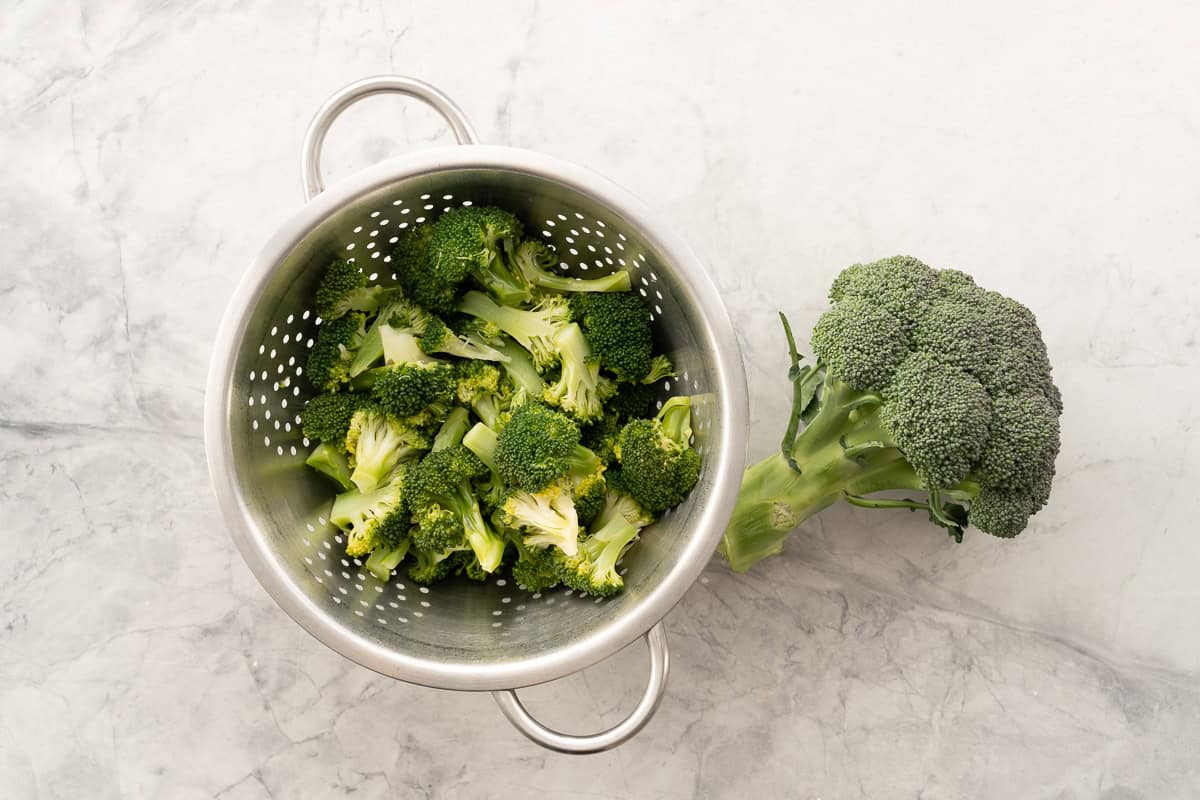 cooked broccoli florets in colander with whole broccoli laid on bench top next to it.