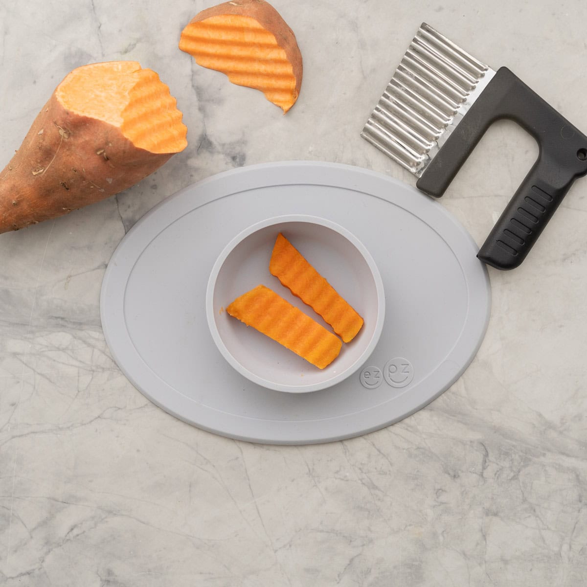 Baby silicone plate on bench top with two wedges of cooked orange kumara. Crinkle cutter and whole kumera next to plate.