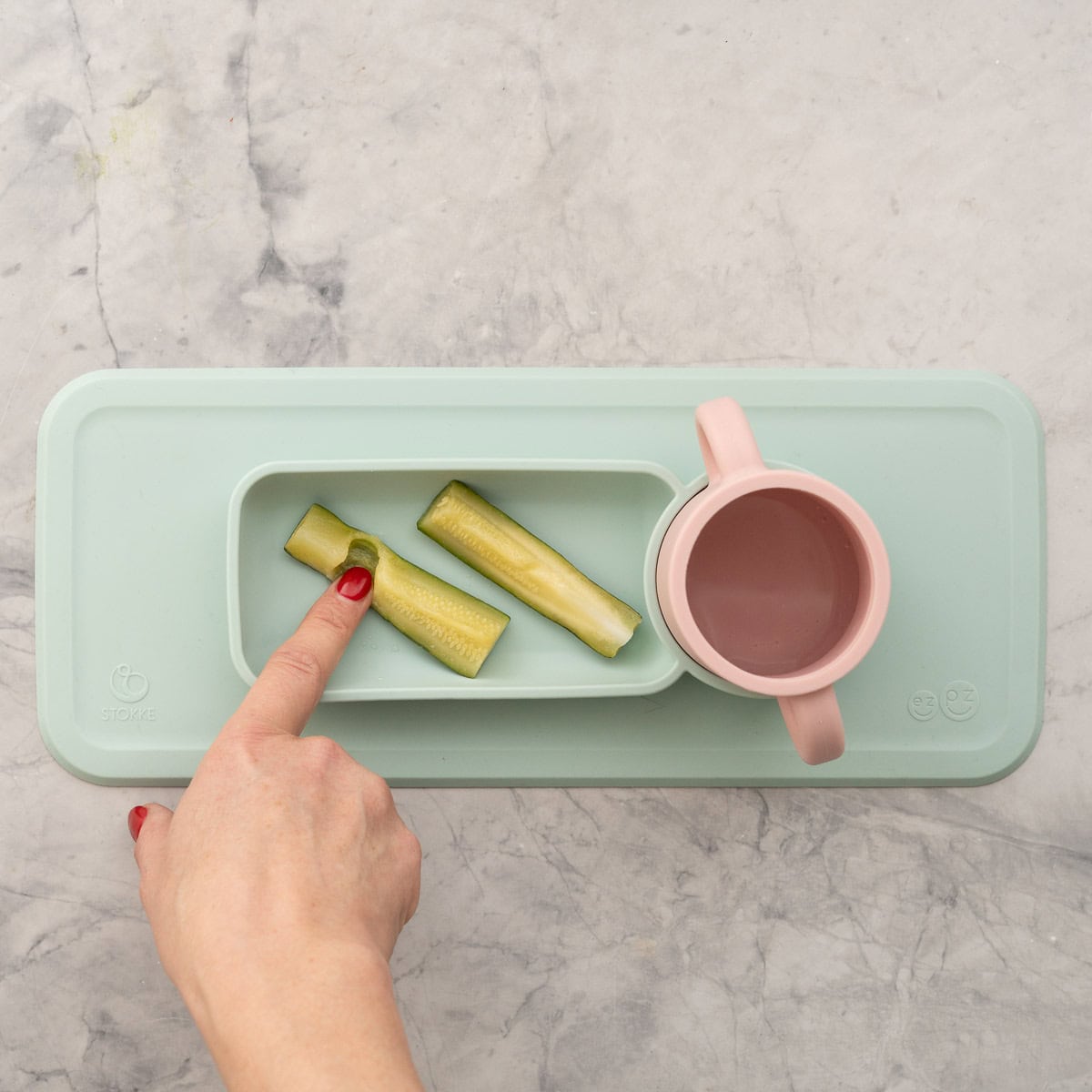 Two slices of zucchini in baby silicone plate with water cup. One finger pressed into a piece showing it is mushy to touch.