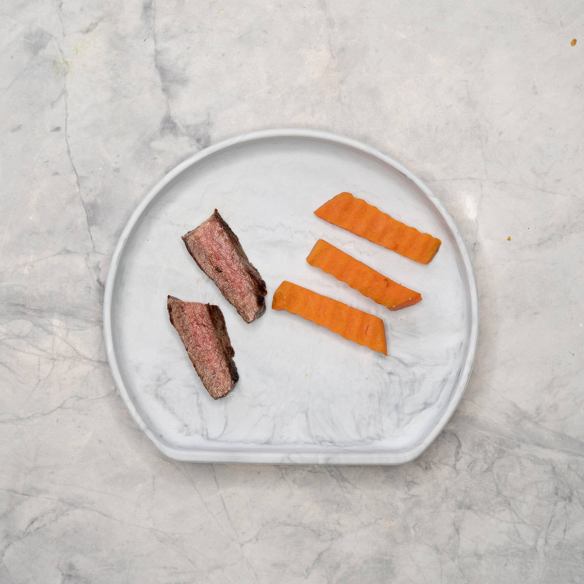 Two slices of cooked steak medium rare, three pieces of cooked kumara cut by crinkle cutters on baby silicone plate.