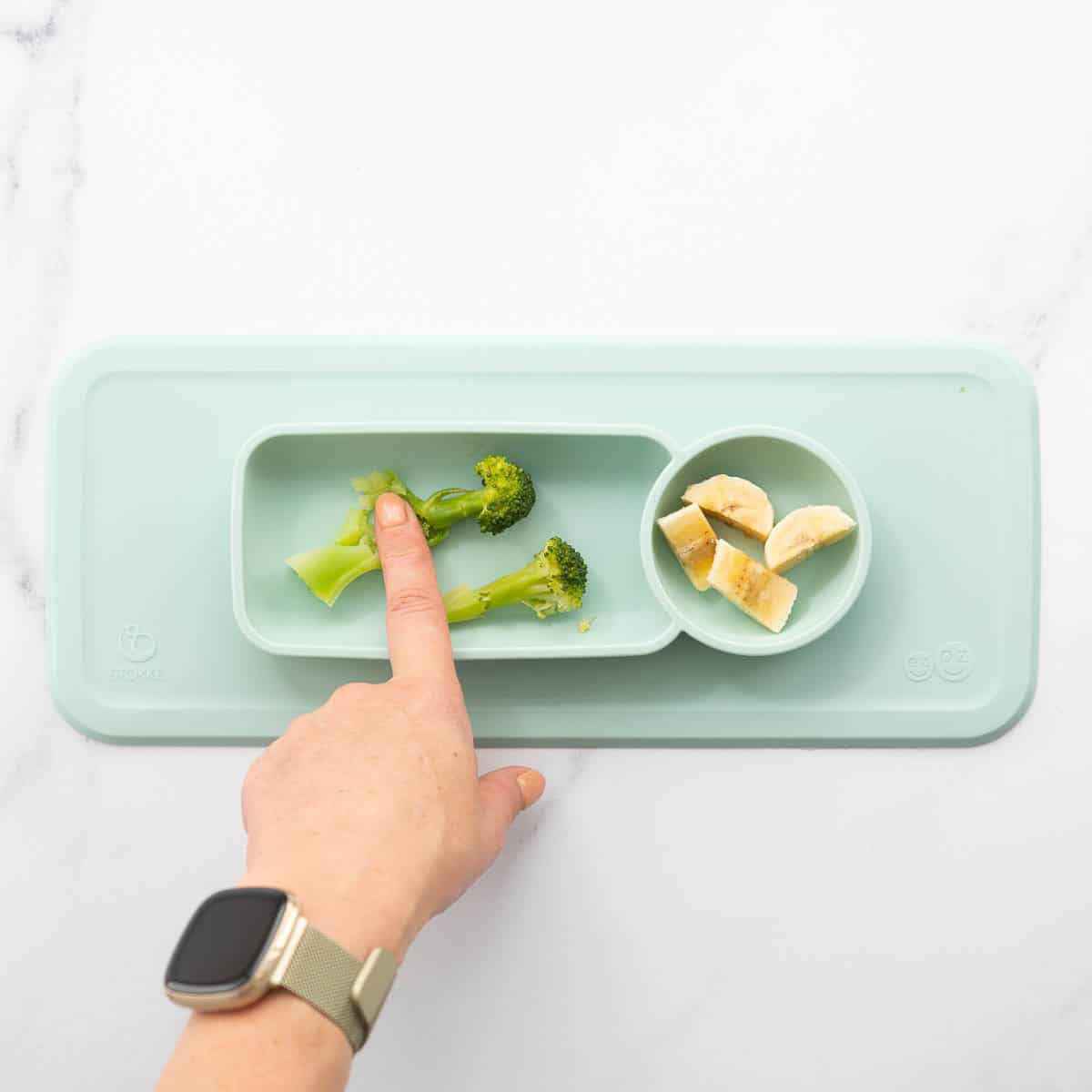 A light blue baby plate of broccoli and banana piece, a finger pushing down on one broccoli floret showing it squishing. 