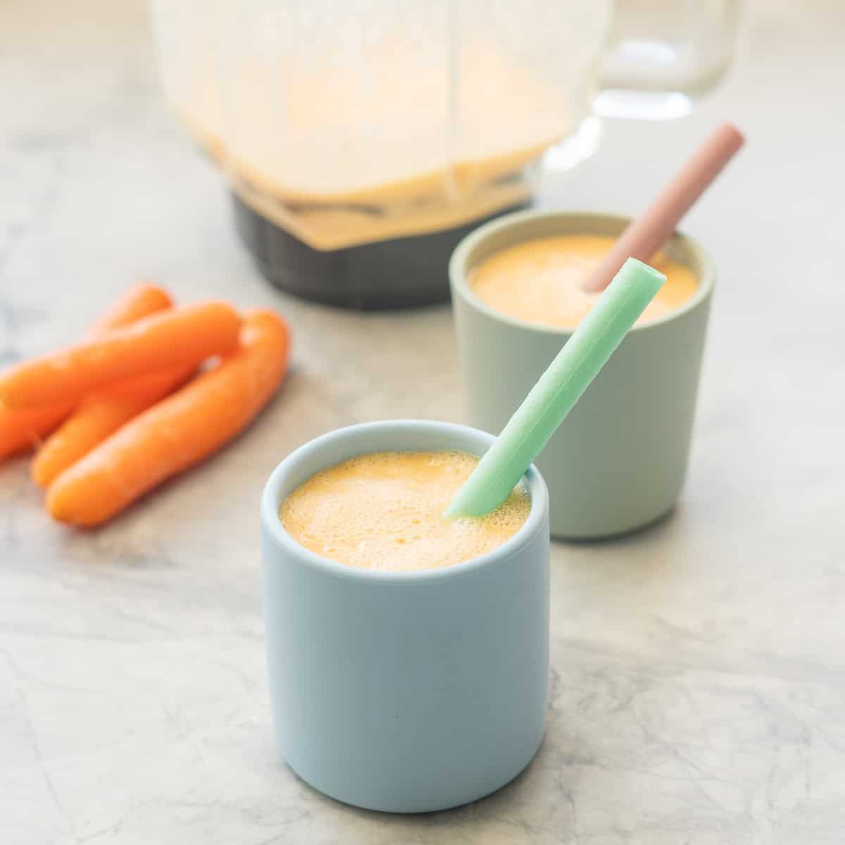 Vibrant orange carrot smoothie in pastel blue and green silicone children cups with colourful straws. Smoothie blender and raw carrots blurred in background.