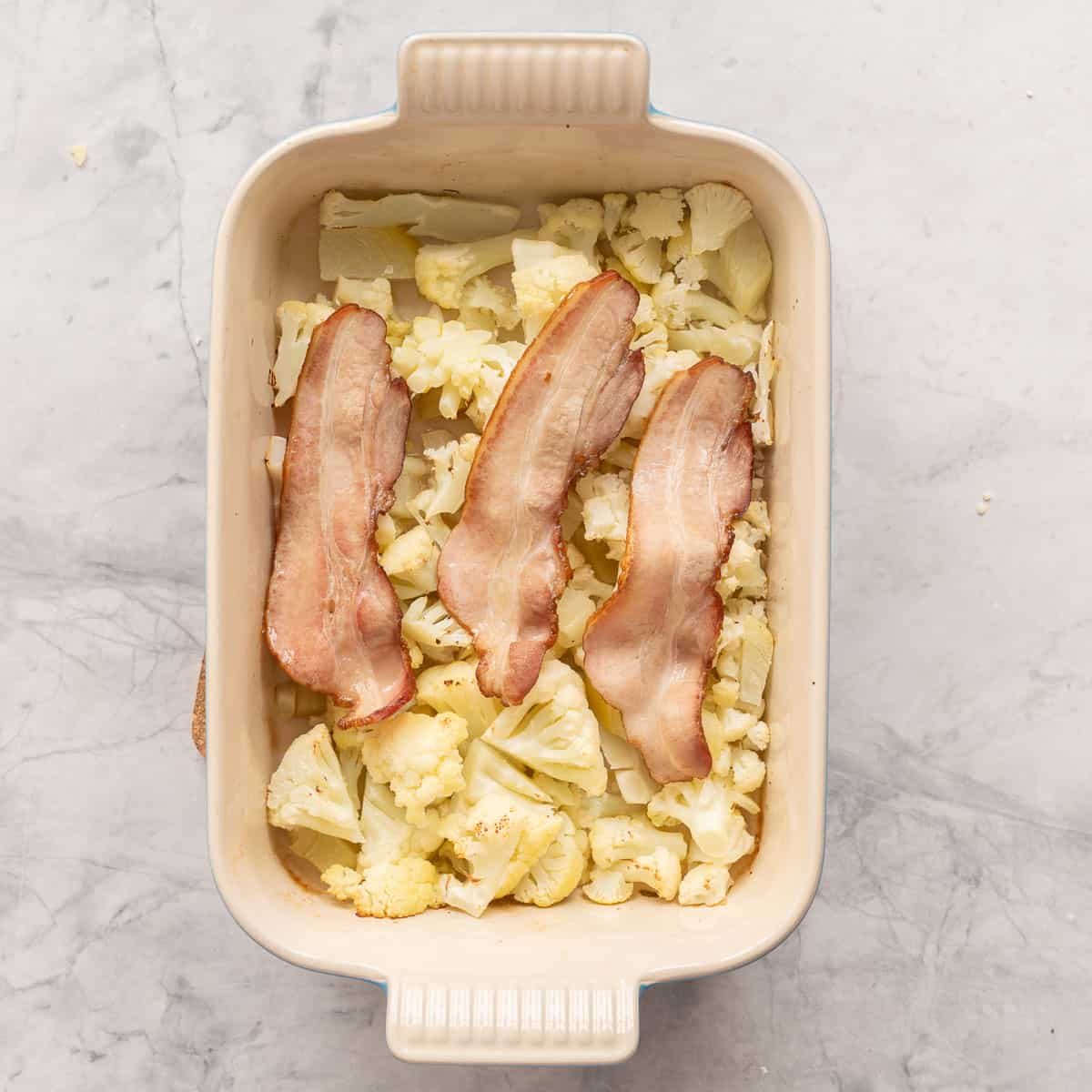 Cauliflower and onion in bottom of baking dish with three pieces of streaky layered bacon on the top.