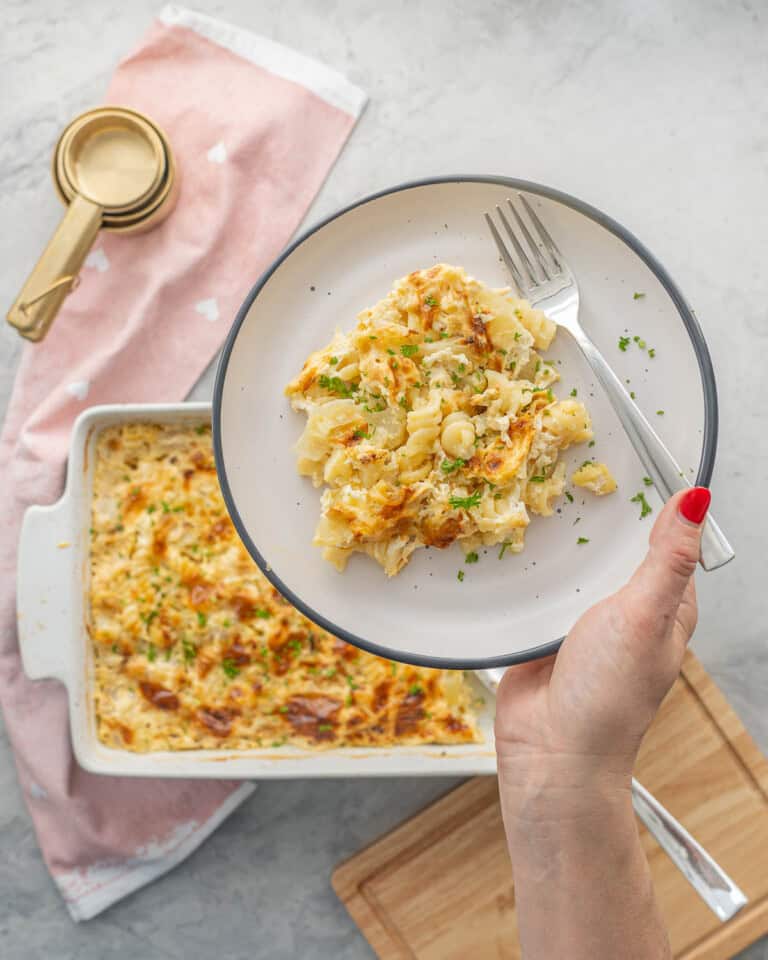 Serving of pasta bake on a dinner plate with fork held up to camera, baking dish with rest blurred in background.