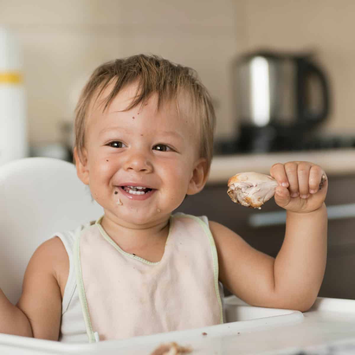 A toddler sitting in a high chair smiling and holding a chicken drumstick. 