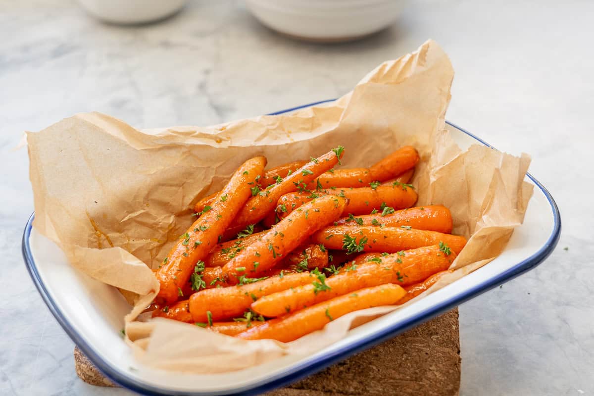 Caramelised glazed carrots placed on brown baking paper in serving dish, garnished with chopped parsley.