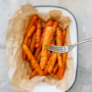 Cooked glazed carrot garnished with chopped parsley being held up by fork, the serving dish with the rest blurred in background.