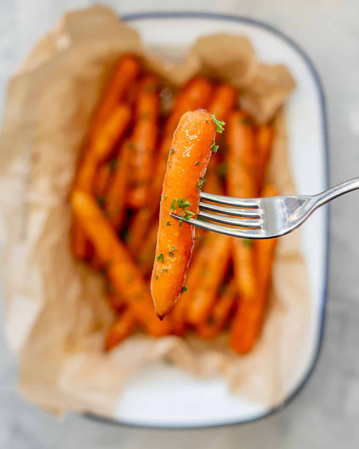 Cooked glazed carrot garnished with chopped parsley being held up by fork, the serving dish with the rest blurred in background.