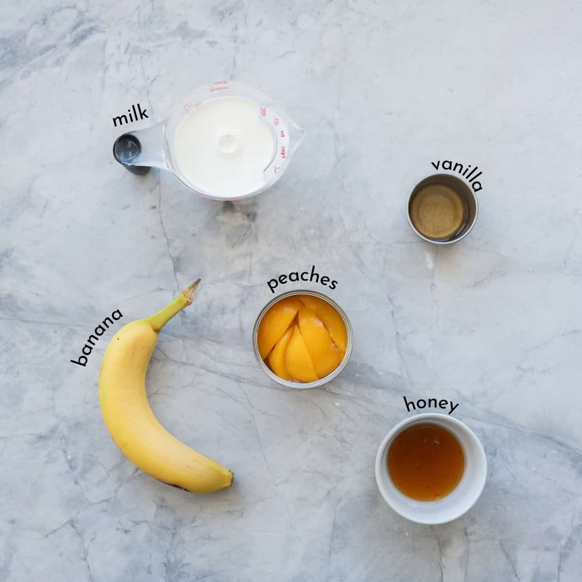 A measuring cup of milk, a can of peaches, banana and small ramekins of honey and vanilla extract on a bench with text overlay. 