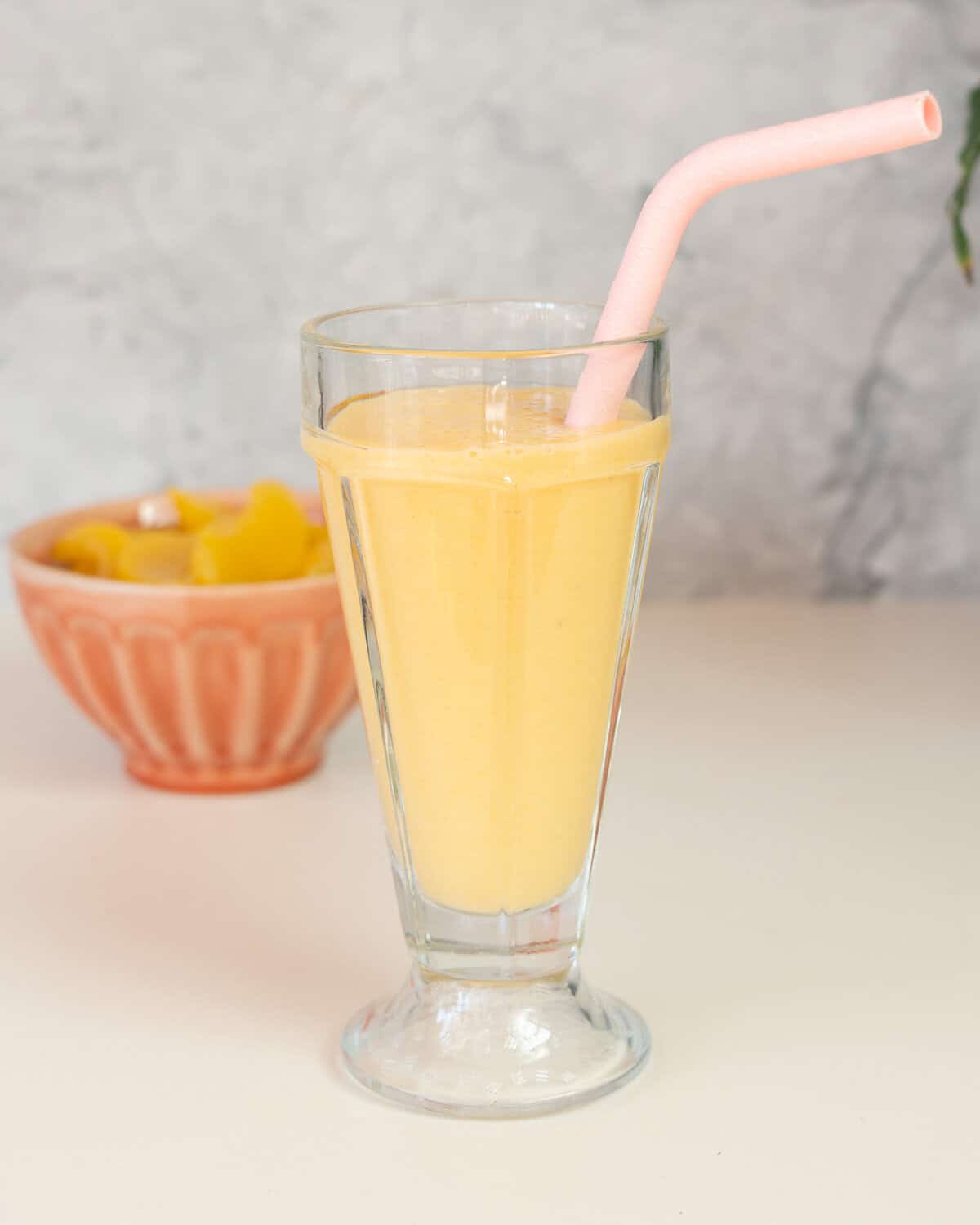 A tall sundae glass filled with a pale yellow smoothie with a pink straw, bowl of peaches in the background.