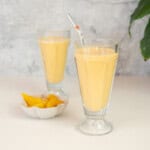 A tall sundae glass filled with a pale yellow smoothie with a pink straw, bowl of peaches in the background.