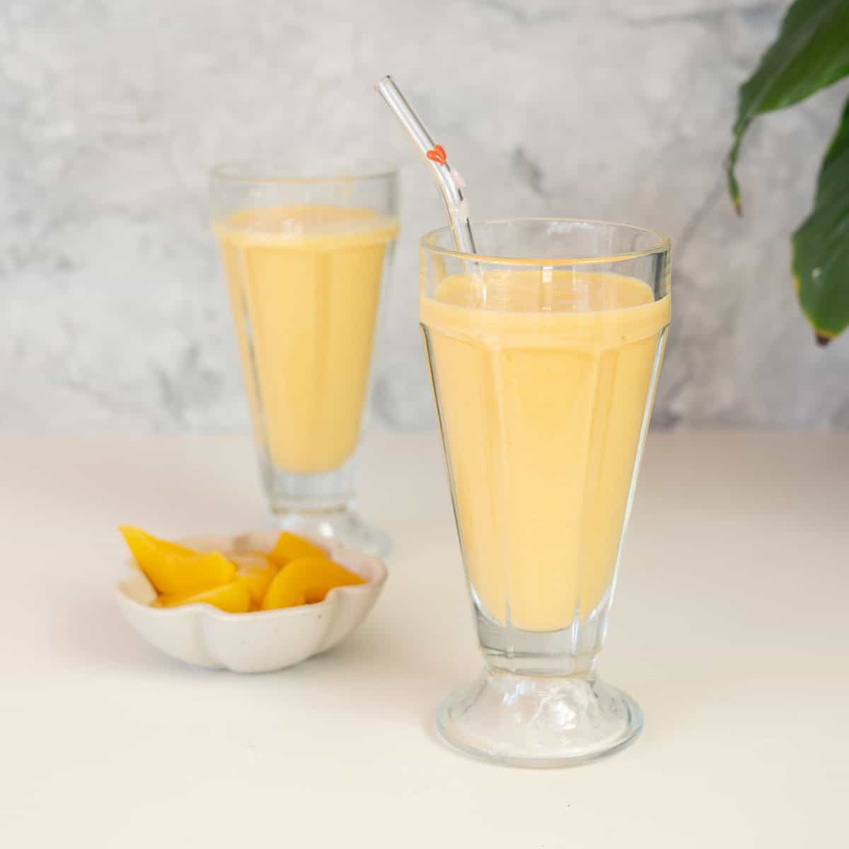 Two tall sundae glasses filled with a pale yellow smoothie with a pink straw, a small bowl of peaches in the background. 