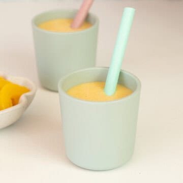 Two small green toddler cups with silicone straws filled with peach smoothie on a white counter top.