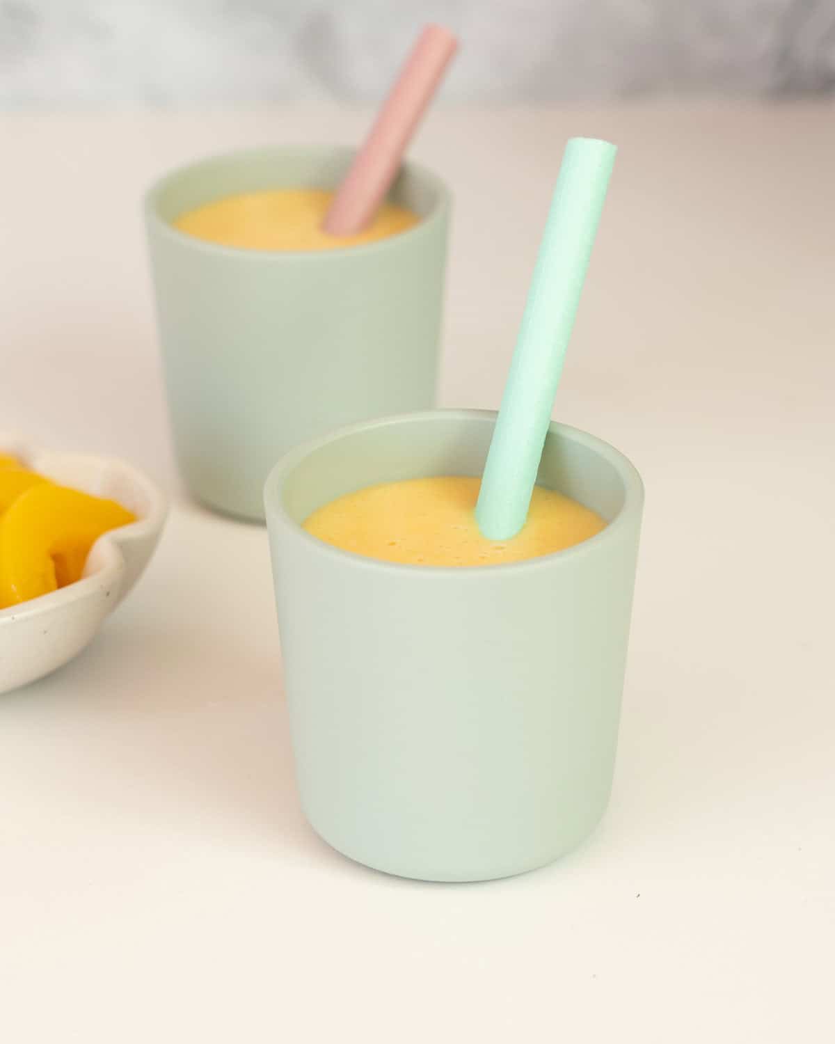 Two small green toddler cups with silicone straws filled with peach smoothie on a white counter top. 