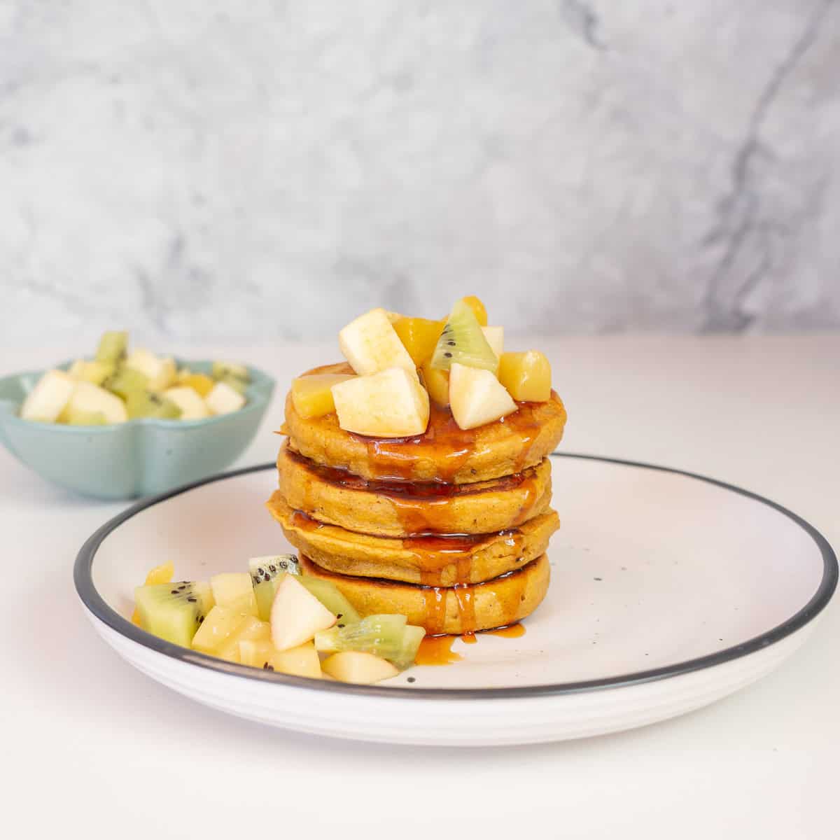 A stack of 5 orange coloured pancakes on a plate topped with fruit salad and drizzled with syrup.