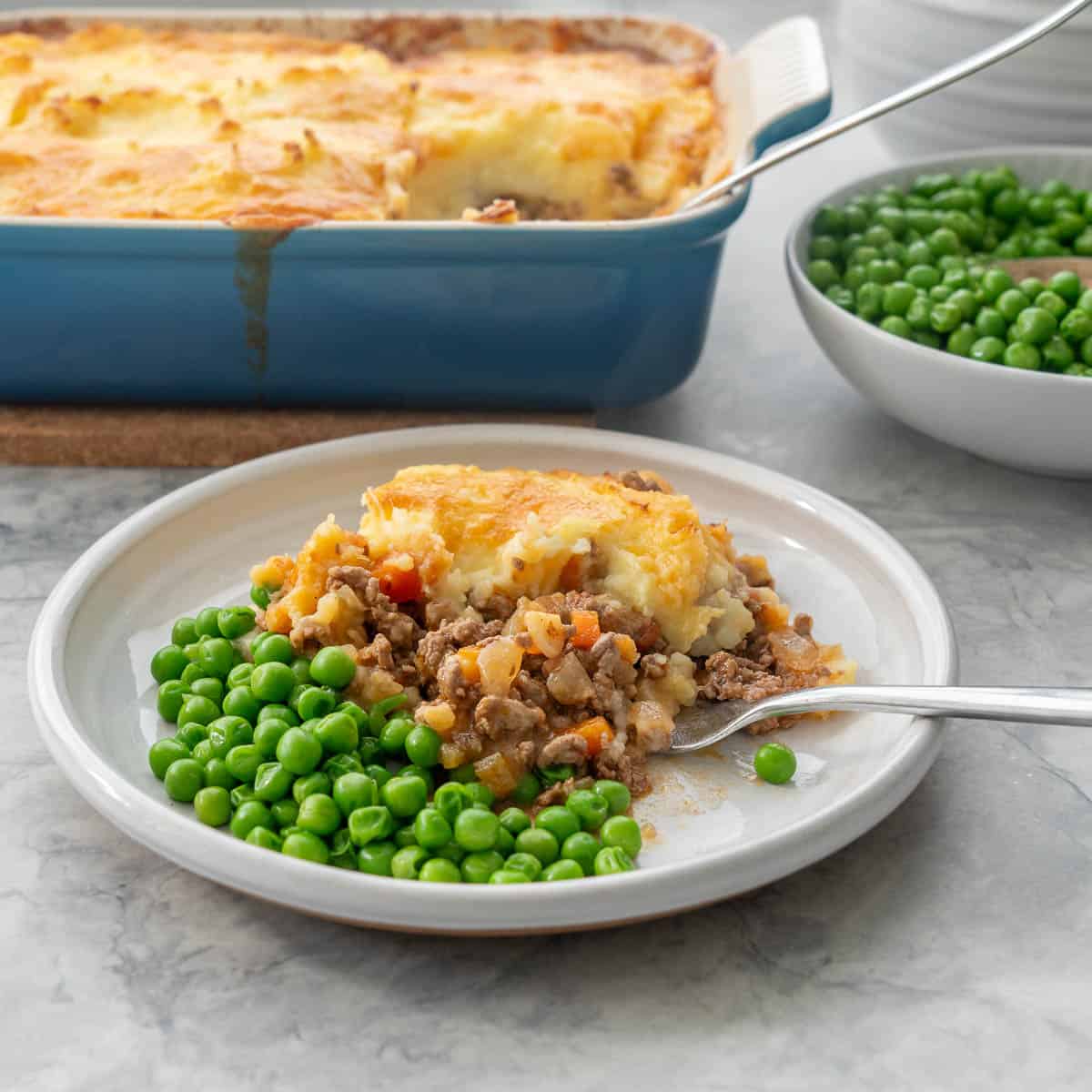 Shepherds pie on dinner plate with serving of peas and fork. Baking dish and bowl of peas blurred in background.