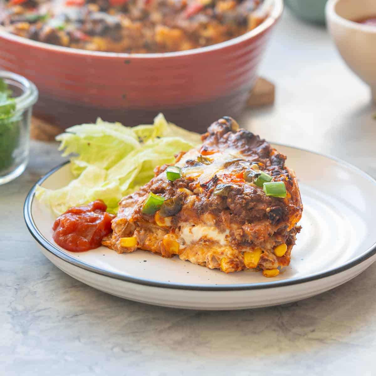 A wedge of Mexican lasagne on a dinner plate, corn kernels. melted cheese, minced beef and sour cream oozing out between the layers.