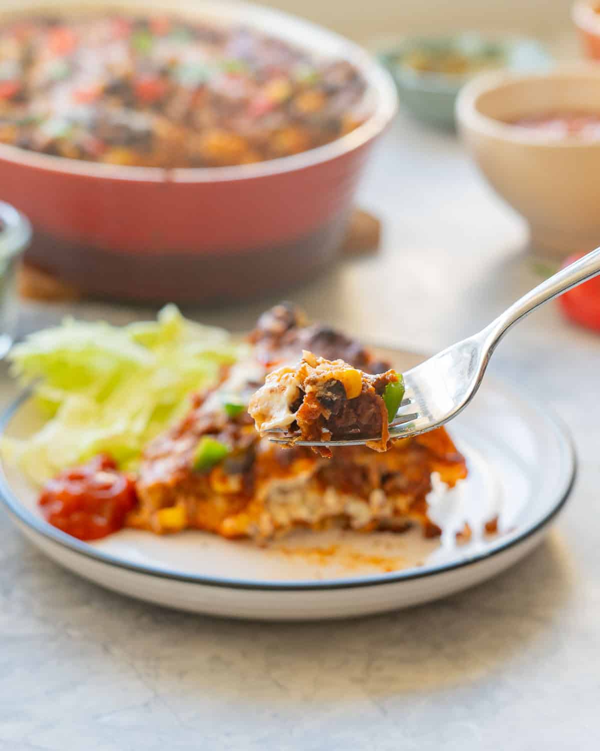 A fork full of Mexican lasagne being lifted up in from to wedge of Mexican lasagne on a dinner plate, corn kernels. melted cheese, minced beef and sour cream oozing out between the layers.