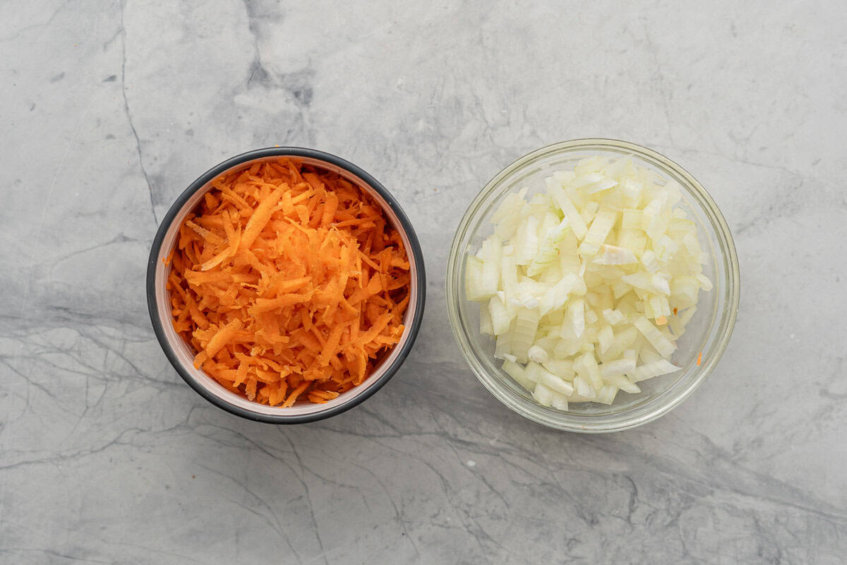 A bowl of grated carrot and a bowl of diced onions on a marble bench top.