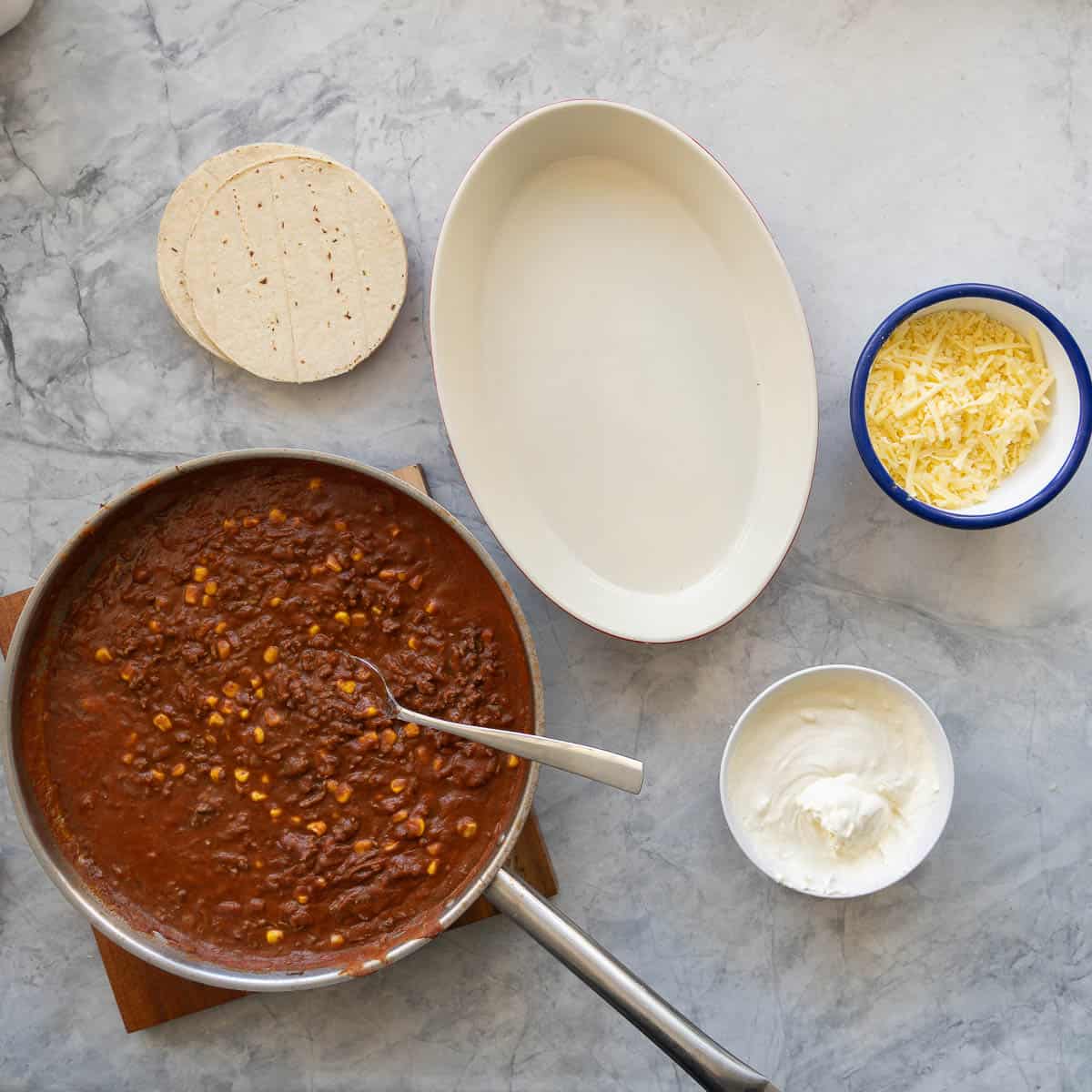 An empty ovel baking dish, next to a large fry pan of mexican mince, a bowl of sour cream, tortillas and a bowl of grated cheese.