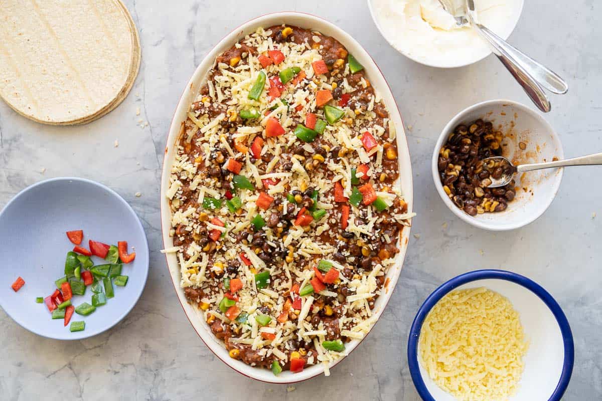 A tortilla casserole ready to go in to the oven, topped with minced beef topped with grated cheese, diced capsicum, black beans and corn kernels.