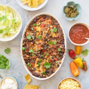 A casserole topped with melted cheese, diced capsicum, corn and beans on a bench top scattered with tortilla chips and coriander leaves.