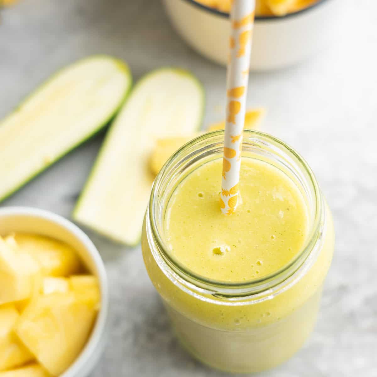 Looking down into a tropical smoothie in tall glass with straw. Chopped pineapple in bowl and sliced zucchini blurred in background.