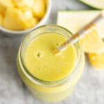 Looking down into a tropical smoothie in tall glass with straw. Chopped pineapple in bowl blurred in background.