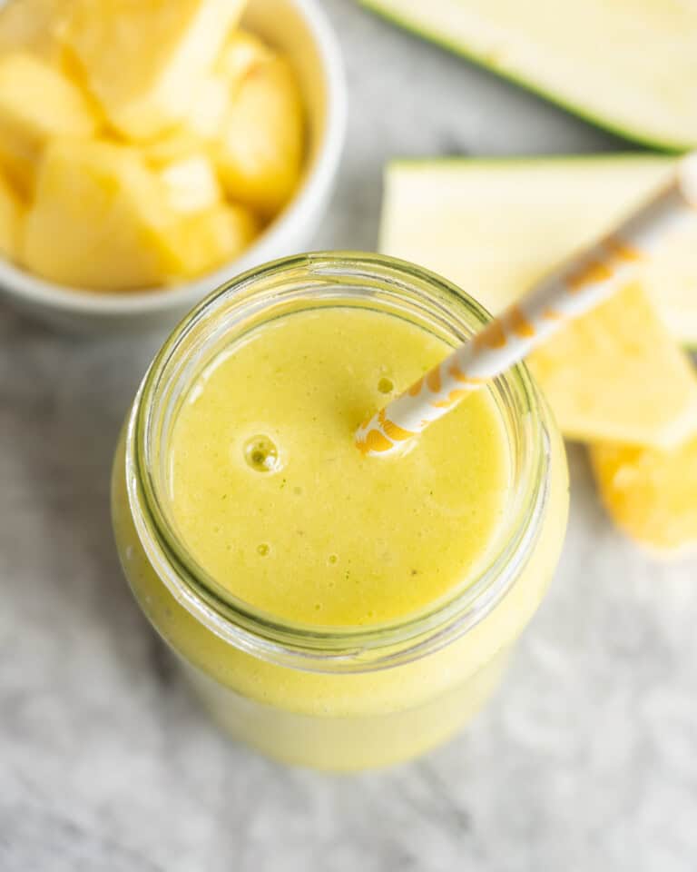 Looking down into a tropical smoothie in tall glass with straw. Chopped pineapple in bowl blurred in background.