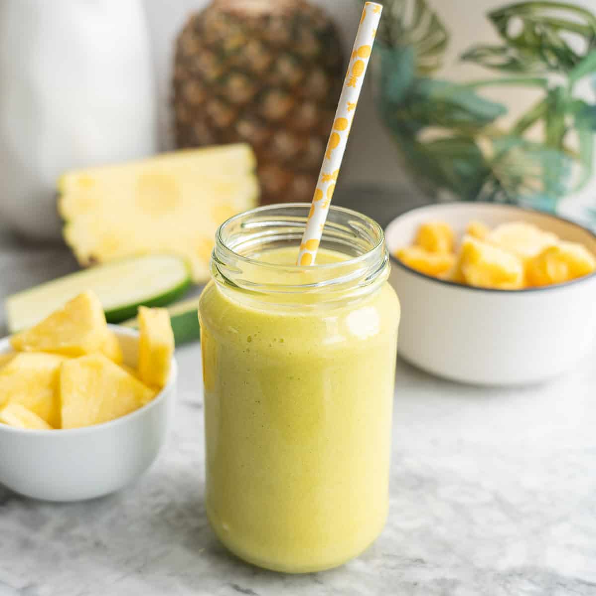 Tropical smoothie in tall glass with straw, chopped pineapple in bowl and whole pineapple blurred in background.