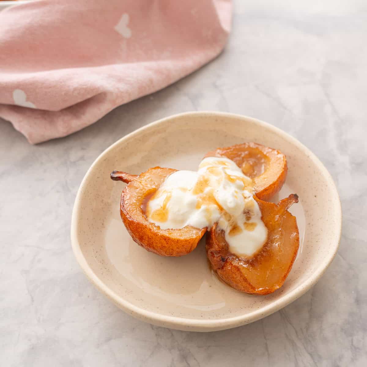 Three caramelised baked pears served on a plate with greek yogurt and a drizzle of maple syrup on top.