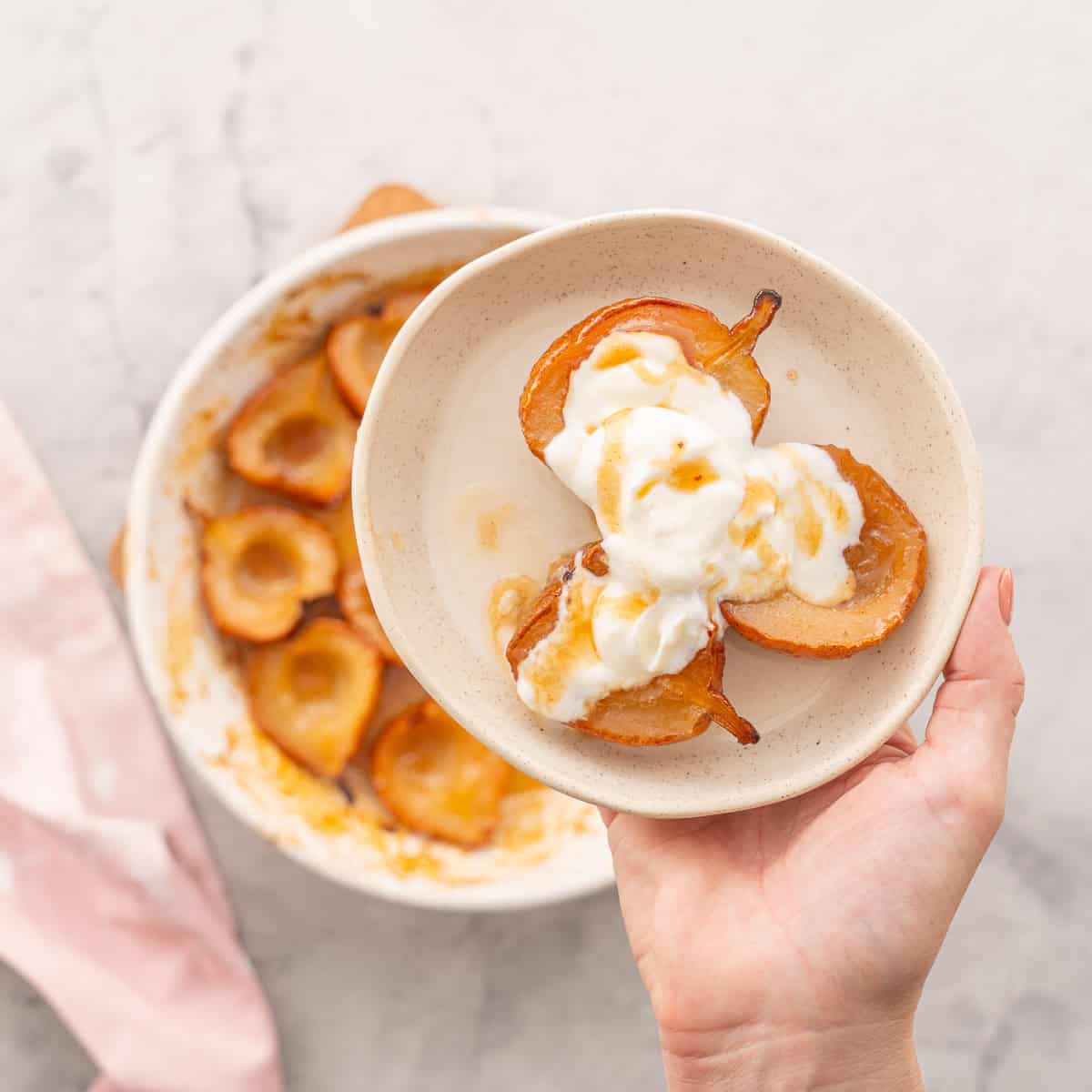 Three caramelised baked pears served on a plate with greek yogurt and a drizzle of maple syrup on top, baking dish with the remaining pears blurred in background.